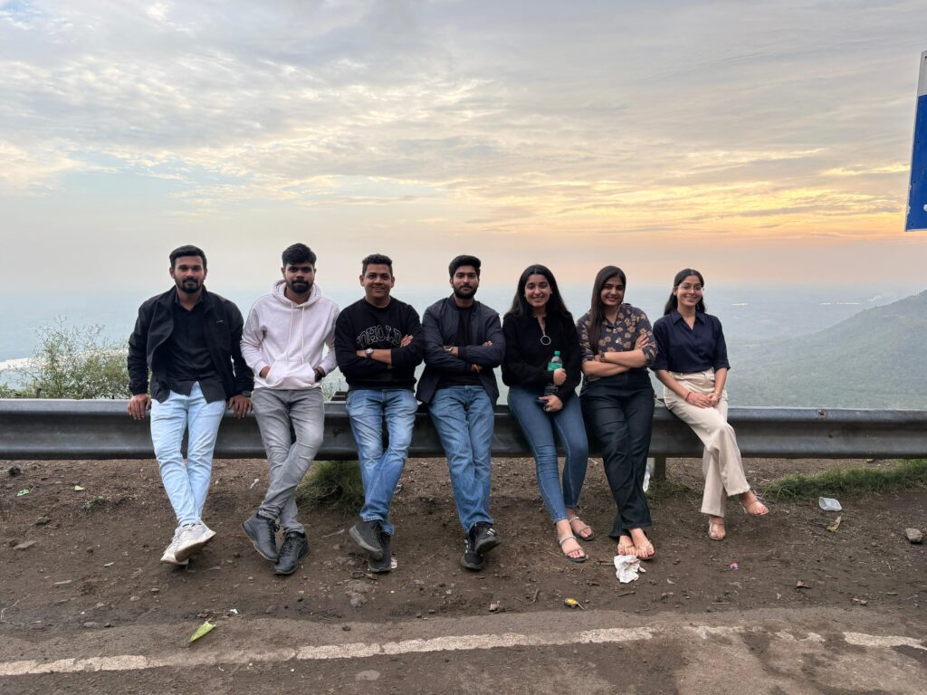 Team outing photo of young professionals sitting on a roadside railing with a scenic mountain view and sunset background.